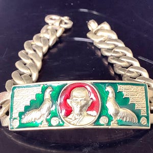 May include: Gold-toned chain bracelet with a rectangular centerpiece. The centerpiece features a central medallion with a figure in red and gold, flanked by two gold bird figures on a green background. The chain links are thick and rounded.