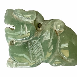 May include: A translucent green carved figurine of a mythical creature. The figurine has a detailed design, with visible features including a head, body, and tail. It has a hole drilled through the top, suggesting it may be a pendant or charm.