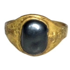 Puede incluir: Un anillo dorado con una piedra ovalada de color gris oscuro en el centro. La banda del anillo tiene una apariencia texturizada, y la piedra es lisa y pulida. El anillo probablemente está hecho de metal y está diseñado para ser usado en un dedo.