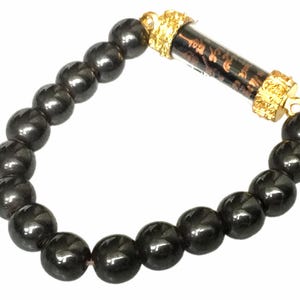 May include: A black beaded bracelet with a gold-colored cylindrical charm. The bracelet features large, round, glossy black beads. The charm is encased in gold-colored metal with ornate detailing. The bracelet is displayed against a white background.