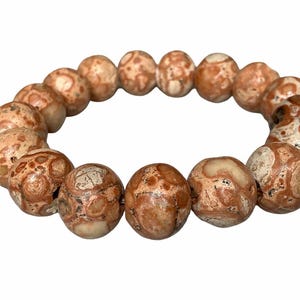 Könnte beinhalten: Ein Armband mit großen, runden Perlen in Braun- und Cremetönen. Die Perlen haben ein gesprenkeltes Muster, das einen natürlichen Look erzeugt. Das Armband ist aufgefädelt und sofort tragbar. Die Perlen haben einen Durchmesser von ca. 2 cm.