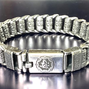 Puede incluir: Pulsera plateada con un diseño detallado y enlazado. La pulsera presenta eslabones rectangulares con patrones grabados y un cierre central con un emblema circular. El estilo sugiere una estética vintage o artesanal, con unos 2 cm de ancho.