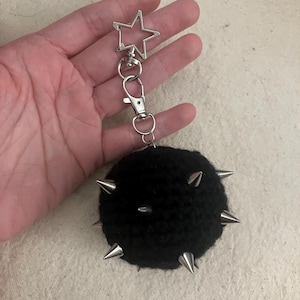 May include: A black, round keychain charm with silver metal spikes and a star-shaped clasp. The charm is made of a fuzzy material and has a silver metal clasp and key ring.