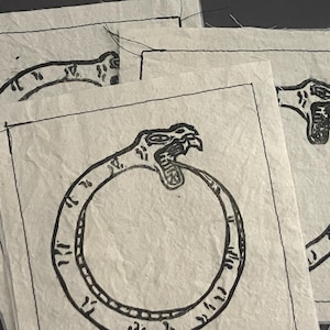 May include: Several handmade paper cards with a black line drawing of an ouroboros, a serpent eating its own tail. The cards have a textured, off-white appearance and are stacked slightly, showing the detailed artwork.