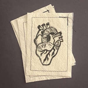 May include: A stack of handmade paper cards featuring a black anatomical heart illustration. The heart design is detailed with veins and chambers, set against a cream-colored background. Each card has a black stitched border.
