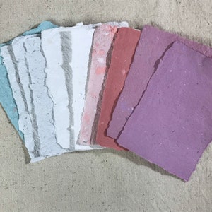May include: A collection of handmade paper sheets in various pastel colors, including blue, white, pink, and purple. The paper has a textured surface and irregular, torn edges, suggesting a craft or art supply.