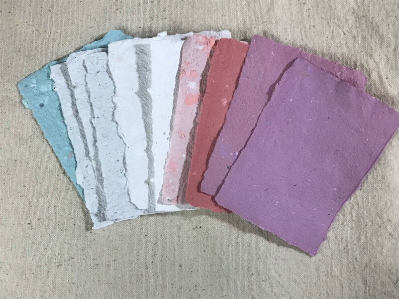May include: A collection of handmade paper sheets in various pastel colors, including blue, white, pink, and purple. The paper has a textured surface and irregular, torn edges, suggesting a craft or art supply.