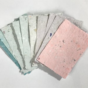 May include: A fan of handmade paper sheets in various pastel colors, including pink, blue, and gray. The paper has a textured surface with flecks of color throughout. The sheets are arranged on a white background.