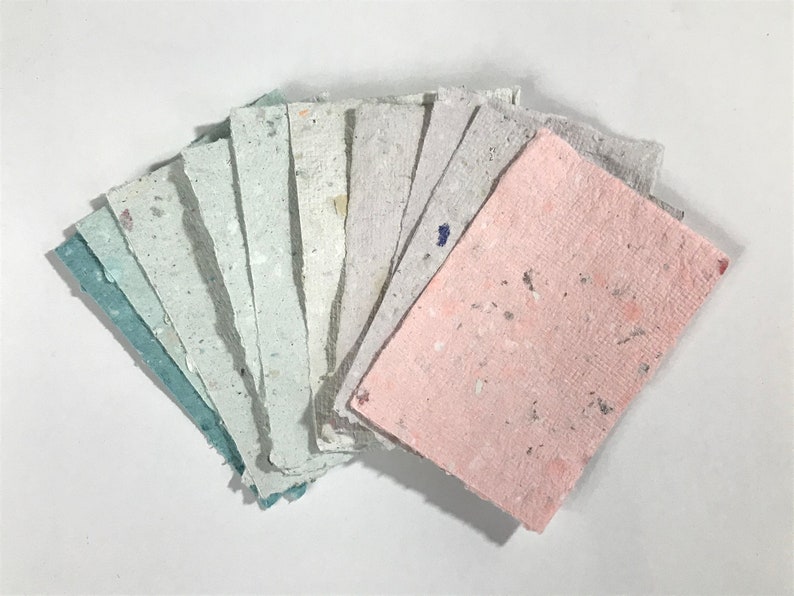 May include: A fan of handmade paper sheets in various pastel colors, including pink, blue, and gray. The paper has a textured surface with flecks of color throughout. The sheets are arranged on a white background.