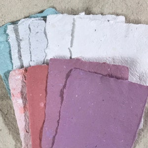 May include: A collection of handmade paper sheets in various pastel colors, including blue, pink, and purple. The paper has a textured, rough edge and is arranged in a fan shape, showcasing the different hues and textures. The background is a neutral canvas.