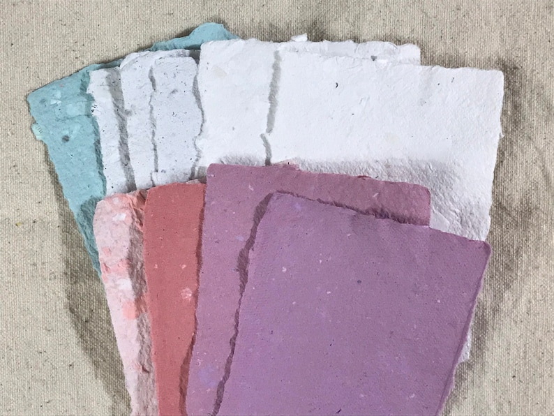 May include: A collection of handmade paper sheets in various pastel colors, including blue, pink, and purple. The paper has a textured, rough edge and is arranged in a fan shape, showcasing the different hues and textures. The background is a neutral canvas.