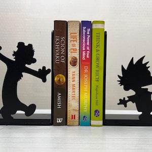 May include: A pair of black metal bookends featuring silhouettes of a cartoon boy and a tiger, supporting four books titled Scion of Ikshvaku, Life of Pi, The Power of Your Subconscious Mind, and Think & Grow Rich, arranged in a vertical row.