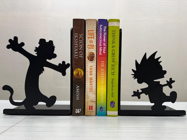 May include: A pair of black metal bookends featuring silhouettes of a cartoon boy and a tiger, supporting four books titled Scion of Ikshvaku, Life of Pi, The Power of Your Subconscious Mind, and Think & Grow Rich, arranged in a vertical row.