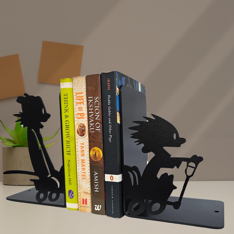 May include: A pair of black metal bookends featuring a silhouette of a cartoon character pulling a wagon. The bookends are positioned on either side of four books titled Think and Grow Rich, Life of Pi, Scion of Ikshvaku, and Hedda Gabler and Other Plays.