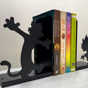 May include: A pair of black metal bookends featuring silhouettes of a cartoon character with spiky hair. The bookends support four books titled Scion of Ikshvaku, Life of Pi, The Power of Your Subconscious Mind, and Think and Grow Rich.