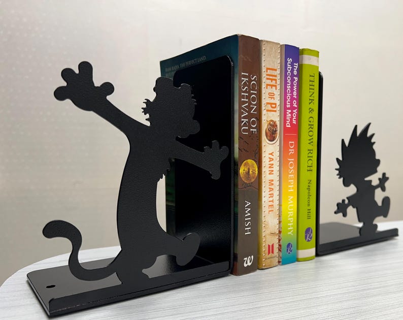 May include: A pair of black metal bookends featuring silhouettes of a cartoon character with spiky hair. The bookends support four books titled Scion of Ikshvaku, Life of Pi, The Power of Your Subconscious Mind, and Think and Grow Rich.