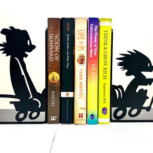May include: A pair of black metal bookends featuring silhouettes of a cartoon character riding in a wagon. The bookends are positioned on either side of five books titled Scion of Ikshvaku, Hedda Gabler and Other Plays, Life of Pi, The Power of Your Subconscious Mind, and Think & Grow Rich.