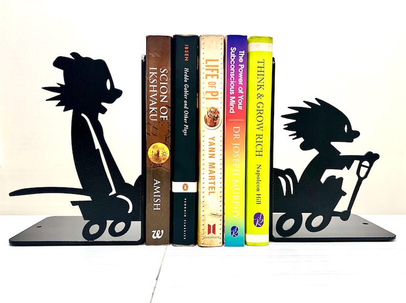 May include: A pair of black metal bookends featuring silhouettes of a cartoon character riding in a wagon. The bookends are positioned on either side of five books titled Scion of Ikshvaku, Hedda Gabler and Other Plays, Life of Pi, The Power of Your Subconscious Mind, and Think & Grow Rich.