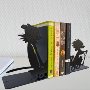 May include: A pair of black metal bookends featuring silhouettes of cartoon characters in wagons. The left bookend shows a character pulling a wagon, while the right shows a character riding in one. They hold four books: Think and Grow Rich, Life of Pi, Scion of Ikshvaku, and Hedda Gabler.