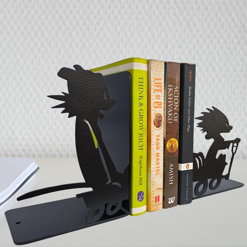 May include: A pair of black metal bookends featuring silhouettes of cartoon characters in wagons. The left bookend shows a character pulling a wagon, while the right shows a character riding in one. They hold four books: Think and Grow Rich, Life of Pi, Scion of Ikshvaku, and Hedda Gabler.