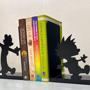 May include: A pair of black metal bookends featuring silhouettes of a cartoon boy and a tiger, holding four books titled Scion of Ikshvaku, Life of Pi, The Power of Your Subconscious Mind, and Think & Grow Rich between them on a flat surface.