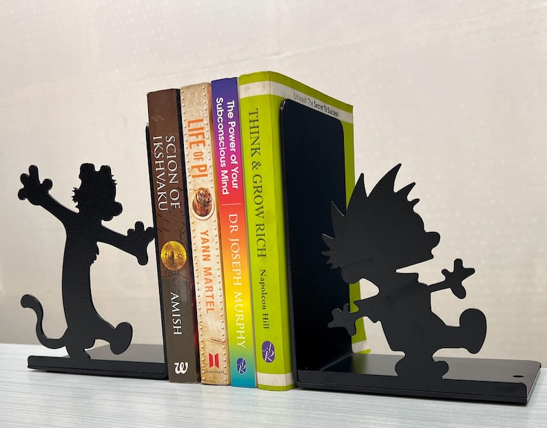 May include: A pair of black metal bookends featuring silhouettes of a cartoon boy and a tiger, holding four books titled Scion of Ikshvaku, Life of Pi, The Power of Your Subconscious Mind, and Think & Grow Rich between them on a flat surface.