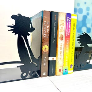 May include: A pair of black metal bookends featuring a silhouette of a cartoon character pushing a small cart. The bookends are positioned on either side of a row of five books with titles including Scion of Ikshvaku, Hedda Gabler, Life of Pi, The Power of Your Subconscious Mind, and Think and Grow Rich.