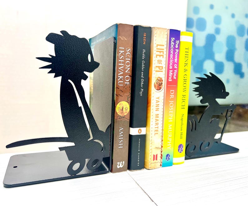 May include: A pair of black metal bookends featuring a silhouette of a cartoon character pushing a small cart. The bookends are positioned on either side of a row of five books with titles including Scion of Ikshvaku, Hedda Gabler, Life of Pi, The Power of Your Subconscious Mind, and Think and Grow Rich.