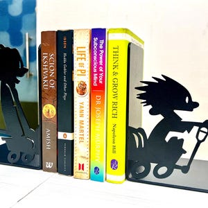 May include: A pair of black metal bookends featuring silhouette cutouts of a character pushing a wagon, holding five books titled Scion of Ikshvaku, Hedda Gabler and Other Plays, Life of Pi, The Power of Your Subconscious Mind, and Think & Grow Rich.