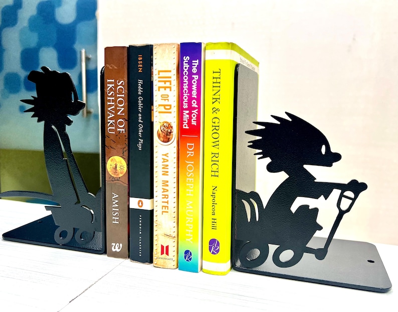 May include: A pair of black metal bookends featuring silhouette cutouts of a character pushing a wagon, holding five books titled Scion of Ikshvaku, Hedda Gabler and Other Plays, Life of Pi, The Power of Your Subconscious Mind, and Think & Grow Rich.