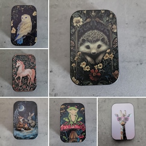 Könnte beinhalten: Eine Sammlung dekorativer rechteckiger Dosen mit verschiedenen Designs. Eine Dose zeigt eine Eule, eine andere einen Igel umgeben von Blumen und eine dritte ein Einhorn. Weitere Dosen zeigen einen Fuchs und ein Kaninchen am Feuer, einen Frosch auf einem Pilz und eine Giraffe mit Sonnenbrille.