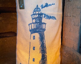 May include: A white canvas bag with a blue embroidered lighthouse design. The bag has a brown leather tag with a white anchor symbol and the letter "T". The bag is lit from inside, creating a warm glow.