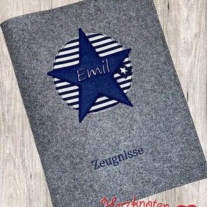 Könnte beinhalten: Grauer Filzordner mit einem blauen Stern und weißen Streifen. Der Stern ist mit dem Namen "Emil" bestickt. Der Ordner trägt die Aufschrift "Zeugnisse" und "Herzknoten" in Rot.