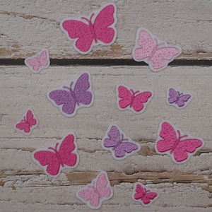 Könnte beinhalten: Eine Ansammlung von gestickten Schmetterlingsaufnähern in verschiedenen Rosa- und Lilatönen. Die Aufnäher haben weiße Ränder und sind auf einer verwitterten, hellen Holzoberfläche angeordnet. Die Schmetterlinge sind unterschiedlich groß.