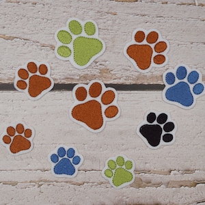 Könnte beinhalten: Eine Auswahl an gestickten Pfoten-Patches in Orange, Grün, Blau und Schwarz. Jeder Patch hat einen weißen Rand und detaillierte Stickereien. Ideal zum Aufnähen auf Kleidung oder Accessoires, die in Zentimetern gemessen werden.