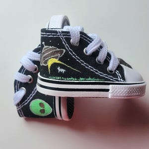 May include: Miniature black canvas sneakers with white laces and soles. The sneakers feature a painted alien and UFO design on the sides. The soles have a black stripe and the toe caps are white.