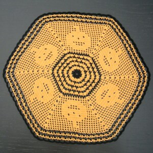 Pode incluir: Uma toalha de mesa hexagonal feita à mão em laranja abóbora com uma borda preta. O centro apresenta um design circular preto, e a toalha de mesa tem um padrão repetido de rostos sorridentes.