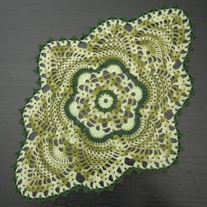 May include: A crocheted doily featuring a complex, symmetrical floral pattern in shades of light green, olive, and dark forest green. The circular center radiates outward into an elongated, scalloped shape with intricate openwork stitching.