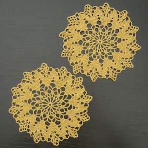 May include: Two crocheted doilies in a light gold colour. The doilies are circular and have a delicate, intricate design.
