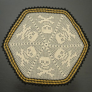 May include: A white crocheted doily with a black and gold border. The doily is hexagonal in shape and has a delicate, intricate design.