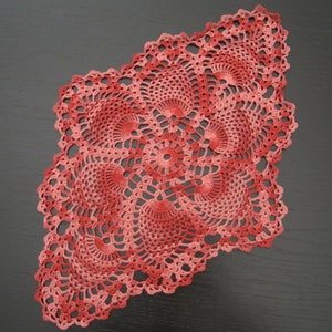 Op de afbeelding: Een delicaat, rood gehaakt tafellaken met een kant-achtig, ingewikkeld ontwerp. Het tafellaken is gemaakt van een fijn garen en heeft een ronde vorm met een centraal punt.
