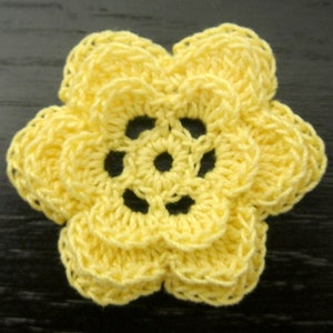 May include: A bright yellow crochet flower applique featuring a layered petal design with a central circular opening. The handcrafted textile piece displays a consistent stitch pattern throughout, creating a dimensional floral shape with six distinct petals.