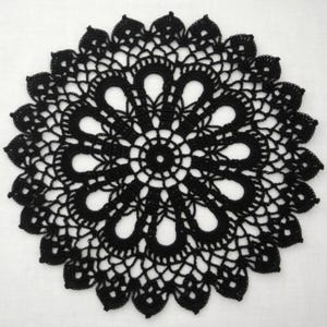 May include: A black, circular doily with an intricate, openwork crochet design. The doily features a central circle surrounded by a series of petal-shaped openings and a scalloped edge. The handmade textile is set against a white background.