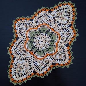 May include: A handmade, crocheted doily with a floral design. The doily features a central pattern in olive green, surrounded by orange and cream-coloured accents. The outer edge is finished with a green scalloped trim, creating a decorative piece.