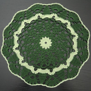 May include: A round, crocheted doily featuring a central light green flower motif surrounded by intricate dark green openwork patterns, finished with a scalloped light green border and a final outer edge of dark green fan-shaped stitches.