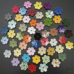Puede incluir: Una colección de pequeñas flores de ganchillo de seis pétalos en una gran variedad de colores sólidos y matizados, incluyendo rojo, azul, amarillo, verde, rosa, morado, naranja, blanco, marrón y gris, cada una con un pequeño orificio central.