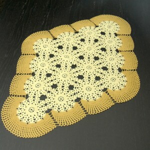 Peut inclure: Un napperon en crochet de forme ovale composé d'un motif central de fleurs jaune pâle interconnectées, entouré d'une bordure festonnée en fil brun doré. Le travail à l'aiguille crée un motif de dentelle délicat et ajouré.