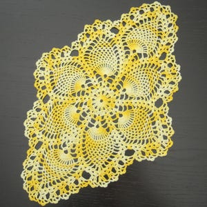 May include: A yellow and white crocheted doily with a delicate, intricate design. The doily is made with a fine thread and has a scalloped edge.