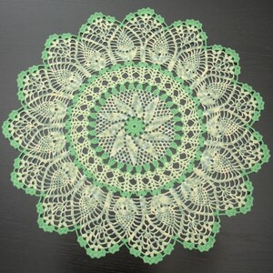 Puede incluir: Un tapete redondo tejido a ganchillo a mano con un complejo patrón calado en tonos variados de verde claro y crema. El diseño irradia desde un motivo floral central, expandiéndose en formas intrincadas similares a piñas con un borde festoneado.