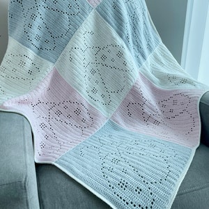 May include: A handmade crochet blanket in a patchwork design. The blanket features squares of white, light pink, and light blue. Each square has a delicate openwork pattern. The blanket is draped over a gray armchair.